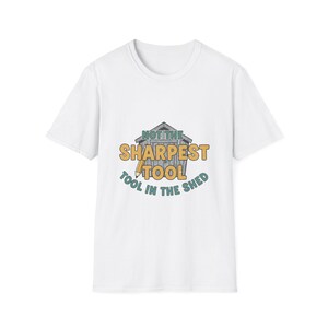 Può includere: T-shirt bianca con la scritta "NOT THE SHARPEST TOOL IN THE SHED". Il design presenta un'illustrazione di un capannone con testo in verde e giallo. Abbigliamento casual.