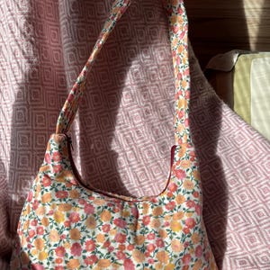Puede incluir: Bolso de hombro de tela con estampado floral. El bolso presenta un diseño curvo y está cubierto de pequeñas flores rosas y naranjas sobre un fondo blanco. La correa y el interior son de color liso. Un accesorio de moda.