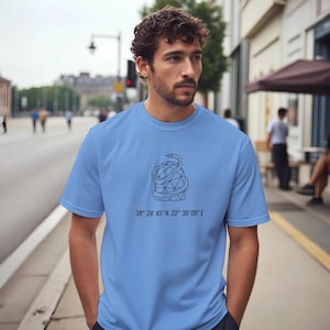 Peut inclure: T-shirt bleu clair avec un motif noir représentant un serpent enroulé autour d'une forme géométrique. Le texte "38° 28' 45" N, 22° 30' 09" E" est imprimé en dessous. Le t-shirt est à manches courtes.