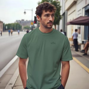 May include: A short-sleeved, olive green t-shirt with a crew neck. The shirt features small black text on the chest. The person is standing on a sidewalk in front of a building.