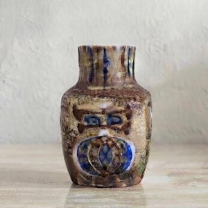 May include: A small ceramic vase with a mottled brown, tan, and cream glaze. The surface features a prominent circular blue and white pattern on the front, accented by dark brown linear markings and a textured, uneven finish around the neck.