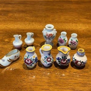Peut inclure: Une collection de onze objets miniatures en céramique comprenant divers petits vases, pichets et un ornement en forme de sabot. Chaque pièce présente des surfaces émaillées blanches décorées de motifs floraux peints à la main et de dorures.