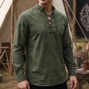 May include: An olive green long-sleeve shirt featuring a mandarin collar and a front placket with a lace-up closure using matching fabric ties. The shirt has long sleeves with buttoned cuffs and a straight hemline, constructed from a solid fabric.