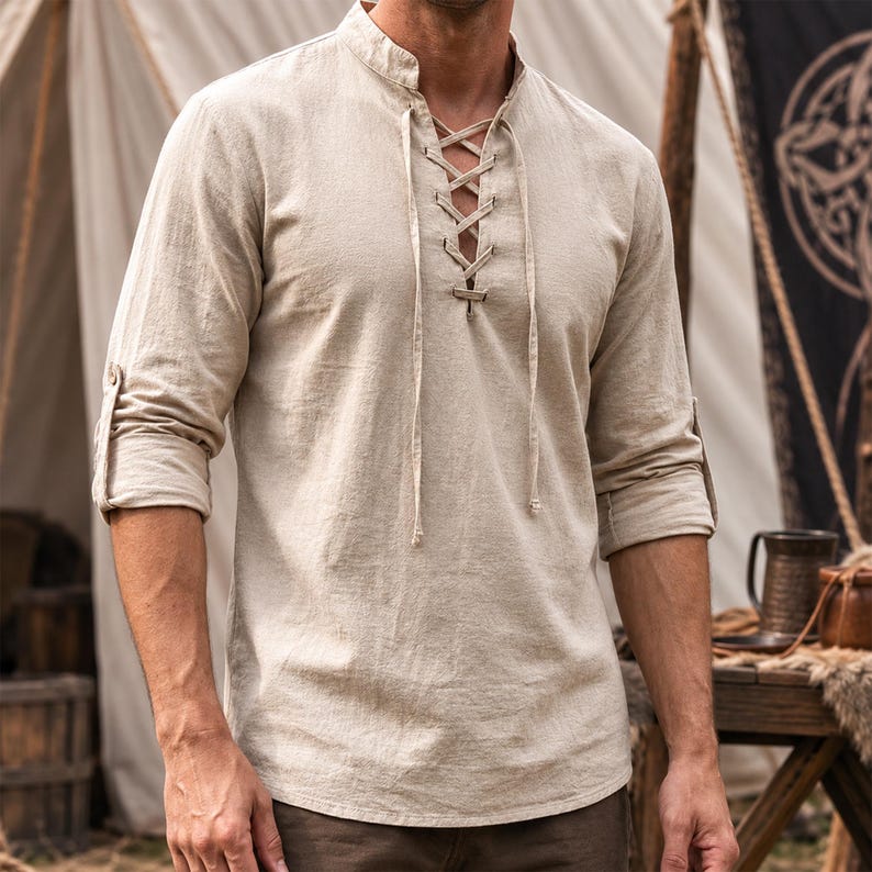 May include: A beige long-sleeve shirt featuring a mandarin collar and a front lace-up placket with matching fabric ties. The sleeves are rolled up to the elbows, and the garment is constructed from a textured fabric with a natural finish.
