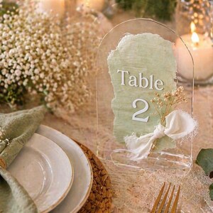 Peut inclure: Numéro de table en acrylique transparent avec un fond peint en vert clair et le texte "Table 2". Le numéro de table est décoré d'un ruban blanc et de fleurs séchées. La table est dressée avec des assiettes blanches, une serviette verte et une fourchette dorée.