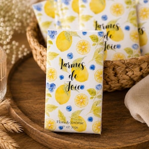 Peut inclure: Boîtes cadeaux rectangulaires blanches avec un motif aquarelle de citrons et de myrtilles. Les boîtes portent le texte "Larmes de Joie" et les noms "Flora & Jérémie" avec la date "27 07 2026". Plusieurs boîtes sont dans un panier en osier.