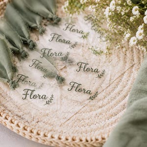 Peut inclure: Une collection d'étiquettes en acrylique transparent, chacune portant le nom Flora imprimé en écriture cursive vert foncé avec un petit motif de feuille. Chaque étiquette est fixée à un ruban en tissu effiloché vert sauge.