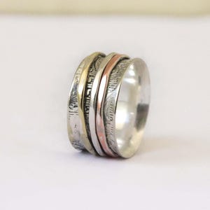 May include: A silver spinner ring with multiple bands. The bands include silver, gold, and rose gold tones. The ring has etched detailing on some of the bands, adding texture and visual interest. The ring is designed to spin.