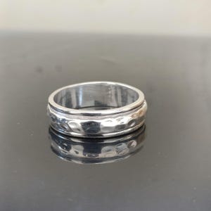 May include: A silver ring with a hammered texture on the outer band. The ring has a smooth inner band and a polished finish. The ring is a simple, yet elegant piece of jewelry. The ring is approximately 0.3 inches wide.