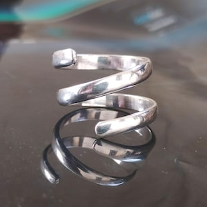 Puede incluir: Un anillo plateado en espiral con un detalle rectangular. El diseño del anillo presenta una banda continua y curvada que se enrolla sobre sí misma, creando una estética moderna y minimalista. El anillo se muestra sobre una superficie reflectante.