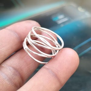 May include: A silver ring with a unique design. The ring features multiple thin, intertwined bands, creating a layered effect. The metal has a bright, reflective finish. The ring is held in a person's hand, with a blurred background.