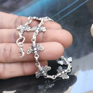 May include: A silver bracelet with a repeating bee motif. The bracelet is composed of linked sections, each with a detailed bee charm. The clasp is visible, and the bracelet is shown against a dark surface. The design is intricate.