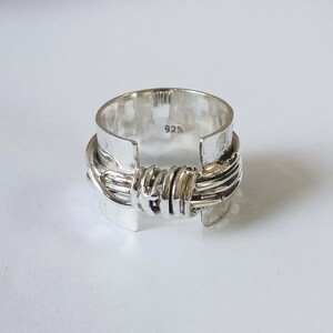 May include: A silver ring with a unique design. The ring features a wide band with a textured, wrapped detail around the center. The metal has a polished finish, and the ring is marked with "925".