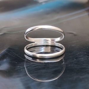 以下が含まれることがあります： 洗練されたモダンなデザインのシルバーダブルバンドリング。リングは、小さなブリッジで接続された2つの平行なバンドを備えており、ユニークで人目を引く外観を作り出しています。磨かれた金属は光を反射し、輝きを増しています。