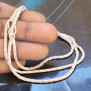 Puede incluir: Un collar de cadena plateada con un diseño tejido. El collar se sostiene en una mano, mostrando su intrincado patrón y el cierre. La cadena parece estar hecha de un metal brillante, que refleja la luz.