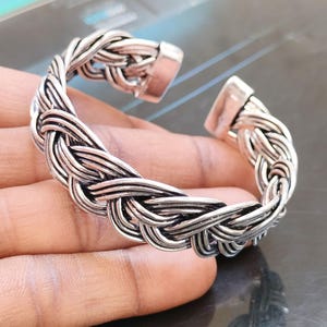 Puede incluir: Brazalete de puño trenzado de color plateado. El brazalete presenta un diseño tejido con un cierre rectangular en cada extremo. El metal tiene un aspecto ligeramente oxidado, lo que le da un aspecto vintage. El brazalete es abierto, lo que facilita su uso.