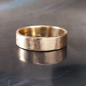 Könnte beinhalten: Ein goldfarbener Ring mit gebürsteter Textur. Der Ring hat ein flaches, breites Band. Der Ring wird auf einer reflektierenden Oberfläche gezeigt. Der Ring ist ein schlichtes, elegantes Schmuckstück. Die Breite des Rings beträgt ca. 0,5 cm.
