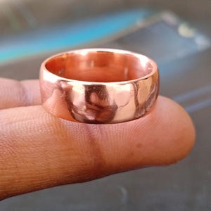 Può includere: Un anello color rame con una fascia larga. L'anello ha una superficie liscia e lucida e riflette la luce, creando riflessi e ombre. L'anello è un design semplice e classico, adatto all'uso quotidiano. L'anello è mostrato su un dito.