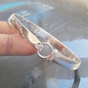 Könnte beinhalten: Ein poliertes silbernes Armreif-Armband mit einem kreisförmigen Verschluss. Das Armband hat ein glattes, abgerundetes Design und einen sicheren Verschluss. Der Verschluss ist ein kleines, kreisförmiges Design, das die beiden Enden des Armbands verbindet.