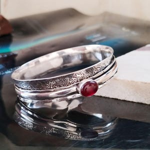 Könnte beinhalten: Ein silberfarbenes Armreif-Armband mit einem eingravierten Blumenmuster und einem roten Edelstein. Das Armband hat ein drehbares Band und eine polierte Oberfläche. Der Edelstein ist oval und in einer Lünettenfassung gefasst.