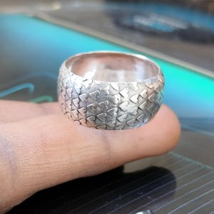 Puede incluir: Un anillo ancho de plata con un patrón de corazones repetidos. El anillo está hecho de un metal brillante y tiene una superficie texturizada. El anillo se muestra en el dedo de una persona.