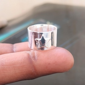 Peut inclure: Une bague argentée avec un motif d'étoile. La bague est large et a une finition polie. L'étoile est en relief sur le devant de la bague. La bague est présentée sur un doigt.