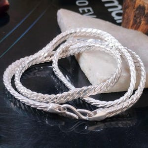 Puede incluir: Un collar de cadena trenzada de color plateado con cierre de mosquetón. El collar se exhibe sobre una superficie negra reflectante con una piedra blanca en el fondo. La cadena tiene una apariencia texturizada y tejida.