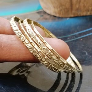 Pode incluir: Quatro pulseiras douradas com padrões florais e de folhas. As pulseiras estão empilhadas, mostrando os detalhes intrincados dos desenhos em relevo. As joias são exibidas em um fundo escuro.