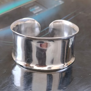 May include: A polished silver cuff bracelet with a wide, curved band. The open design allows for easy wear. The bracelet reflects light, creating a bright, shiny appearance. A simple, elegant piece of jewellery.
