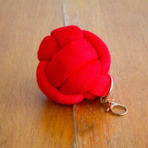 May include: A bright red, velvet-like knot-shaped keychain charm. The charm has a gold-colored clasp for attaching to keys or a bag. The background is a light brown wooden surface.