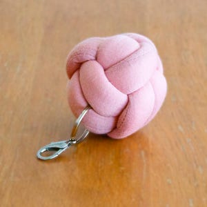 May include: A light pink, knotted fabric keychain with a silver-colored clasp and keyring. The knot is spherical and made of soft material. The keychain rests on a wooden surface.