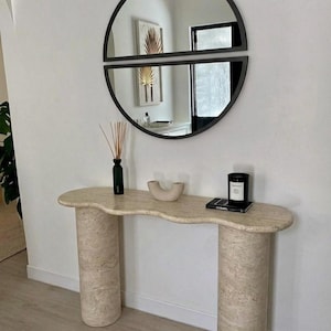 Könnte beinhalten: Eine beige Travertin-Konsole mit gewellter Oberseite und zwei zylindrischen Beinen. Der Tisch ist mit einer schwarzen Kerze, einer kleinen beigen Keramikskulptur, einer grünen Vase und einem Buch dekoriert. Über dem Tisch hängt ein schwarzer, halbkreisförmiger Spiegel.