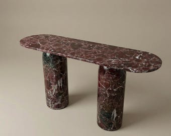 May include: A contemporary console table with a burgundy marble top and two cylindrical supports. The marble displays a striking pattern of dark red, white, and green veining. The table's design is minimalist and elegant, suitable for various interior styles.