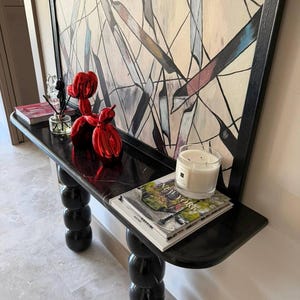 Puede incluir: Una consola de mármol negro brillante, con una escultura de perro globo rojo, una vela y revistas. La mesa tiene un diseño único de patas esféricas apiladas y está debajo de una gran pintura abstracta.