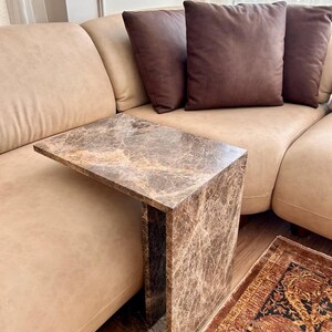 May include: A brown marble-look side table with a rectangular top and a single supporting side. The table is positioned next to a beige sofa with brown throw pillows. A patterned rug is visible on the floor.