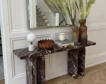 May include: A contemporary console table with a red marble top and base. It features a white globe lamp, a woven wooden bowl, books, a candle, and vases with greenery. A mirror reflects a staircase.