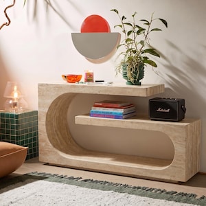 Op de afbeelding: Een beige travertijn console tafel met een C-vormig ontwerp en een grote open ovale uitsparing. Op het tafelblad staan een stapel boeken, een kleine oranje glazen schaal, een kaars en een zwarte Marshall-luidspreker met een plant.