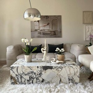 Può includere: Un soggiorno con un tavolino in marmo con venature bianche e nere. Un divano beige con cuscini neri è dietro il tavolo. Gli elementi decorativi includono un candelabro bianco, orchidee in vaso e una lampada a sospensione argentata.