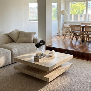 May include: A square, two-tiered coffee table made of light beige travertine stone with a porous, natural texture. The table features a thick, flat top surface and a matching lower shelf, both with clean, sharp edges, resting on a central base.