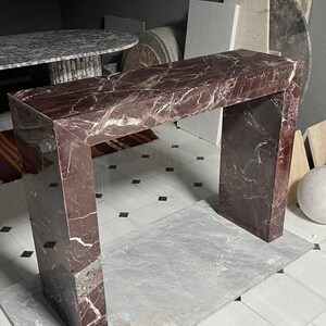 May include: A rectangular marble table with a deep red hue and white veining. The table has a modern, minimalist design with a rectangular top and two supporting legs. The table is sitting on a light gray surface.
