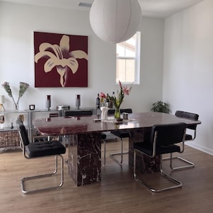 May include: A dining room featuring a large rectangular table with a dark red marble top and base, surrounded by four black upholstered chairs with chrome cantilever frames. A white paper globe pendant light hangs above the table.