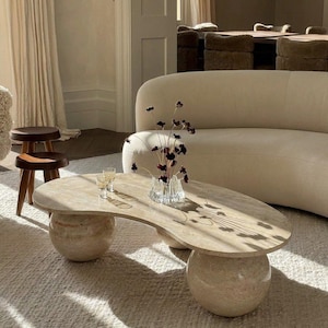 Può includere: Un tavolino da caffè a forma di rene realizzato in pietra travertino beige con una superficie liscia e lucida. Il tavolo è sostenuto da due grandi basi sferiche in travertino. Sopra è posizionato un vaso in vetro trasparente scanalato con fiori secchi scuri.