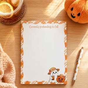 May include: A rectangular notepad with a white center and an orange and white plaid border. The text "Currently pretending its Fall" is printed at the top. A cartoon ghost wearing a straw hat and holding an iced drink sits next to a jack-o'-lantern.