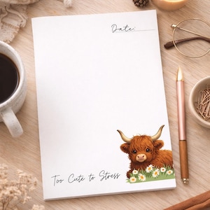 May include: White notepad with a watercolor illustration of a Highland cow and daisies. The notepad has the text "Too Cute to Stress" and a space for the date. A pen, candle, and coffee cup are also visible.