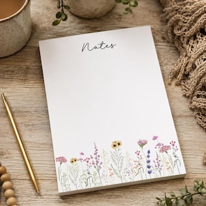 May include: A white rectangular notepad featuring the word Notes in black cursive script at the top and a colorful illustration of various wildflowers in shades of pink, yellow, purple, and green printed along the bottom edge of the paper.