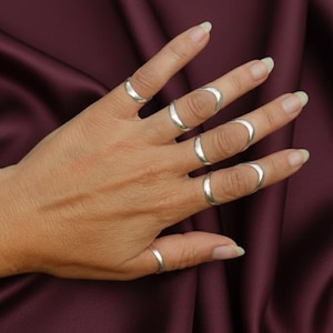 Peut inclure: Une main ornée de plusieurs bagues en argent. Les bagues présentent différents designs, certaines encerclant entièrement le doigt, d'autres formant une boucle partielle. Le fond est un tissu bordeaux foncé, créant un contraste avec le métal brillant.