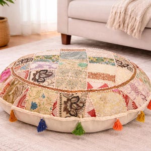 Pode incluir: Um pufe redondo com um design de patchwork em várias cores, incluindo bege, rosa e azul. A almofada tem uma base creme e é adornada com borlas coloridas. Esta almofada decorativa é adequada para uso interno.