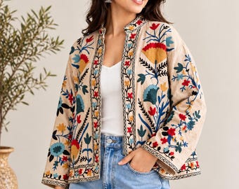 May include: A beige open-front jacket featuring vibrant floral embroidery in shades of red, yellow, and blue. The garment has long sleeves, a mandarin-style collar, and black stitched trim along the edges, worn over a white top and blue jeans.