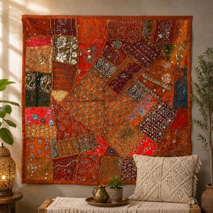 Op de afbeelding: Een vierkant wandkleed bestaande uit een patchwork van oranje, rode en bruine stoffen panelen, versierd met ingewikkeld borduurwerk, metalen lovertjes en kleine spiegeltjes. Het textiel toont een verscheidenheid aan patronen.
