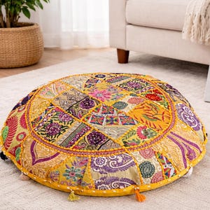 May include: A circular floor cushion with a patchwork design in yellow, purple, and pink. The cushion has a yellow border with small tassels. A painting of white flowers is in the background.